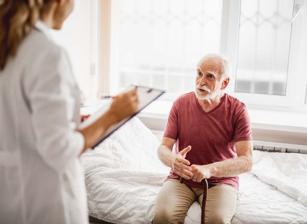 Hospital Indemnity Plans - Senior Male Patient with Cane Talking about His Health with a Female Physician Who is Taking Notes in the Patient’s Hospital Room