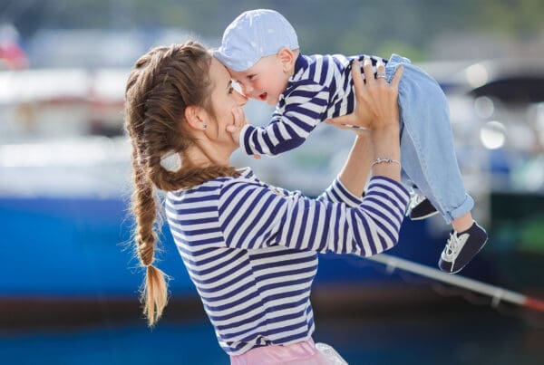 About Our Agency - Cheerful Young Mom Holding her Baby Son Up in the Air While Standing Next to Boats